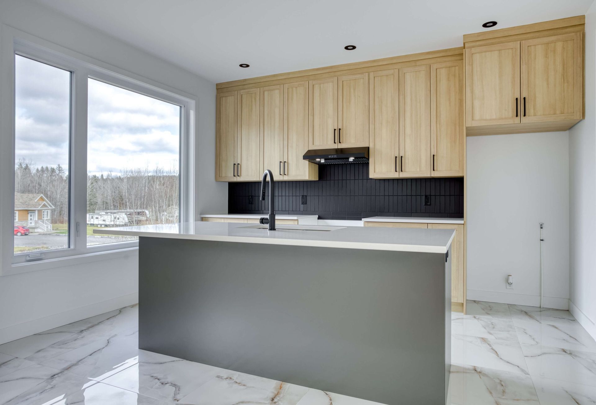 Cuisine moderne avec placards en bois clair, îlot gris et grande fenêtre.