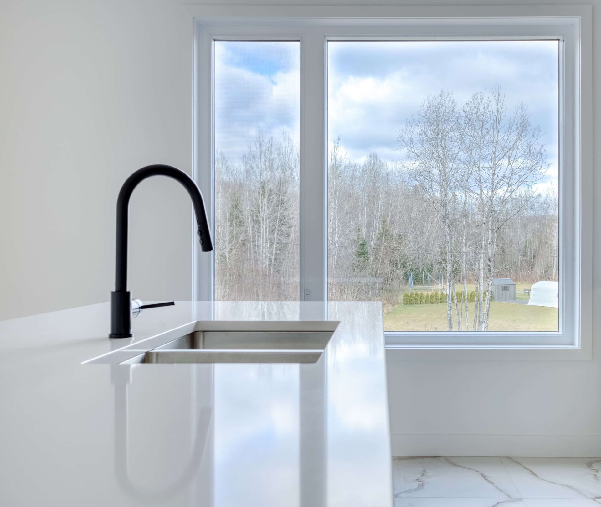 Robinet noir au-dessus d'un évier en acier inoxydable sur un comptoir blanc, avec une fenêtre donnant sur les arbres.