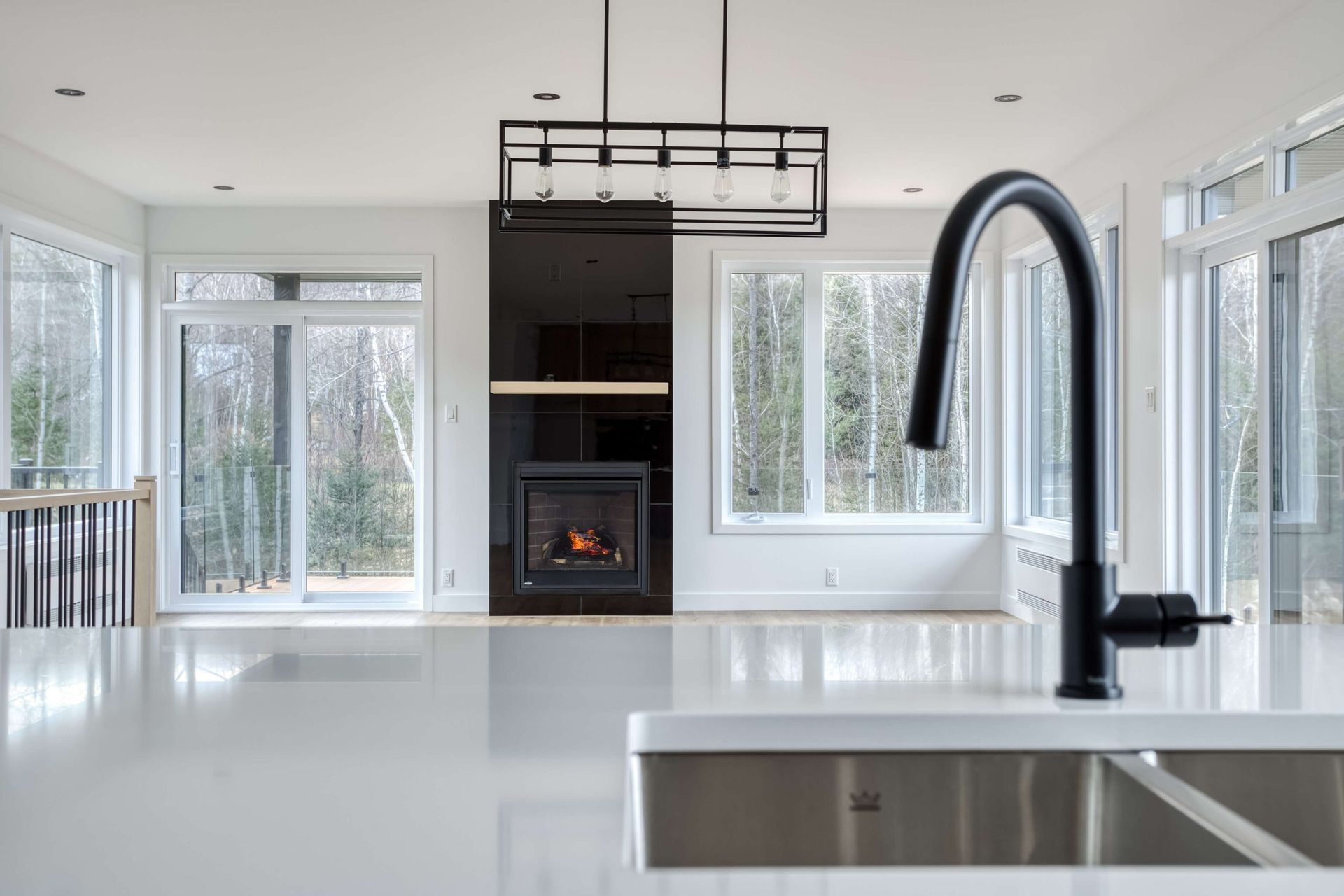 Robinet noir au-dessus d'un plan de travail blanc dans une cuisine moderne. Cheminée et grandes fenêtres en arrière-plan.