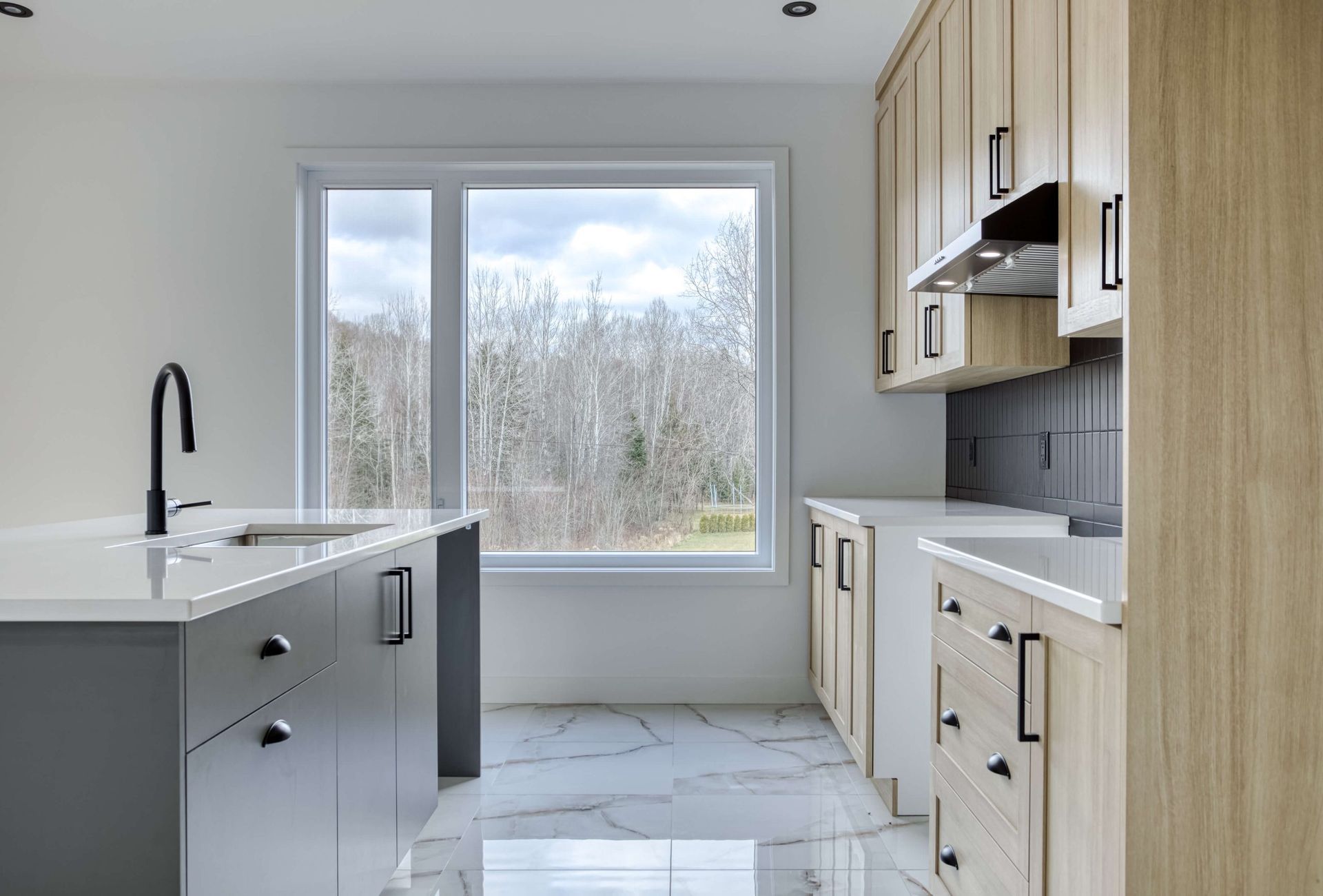 Cuisine moderne avec îlot gris, placards en bois clair et grande fenêtre donnant sur les arbres.