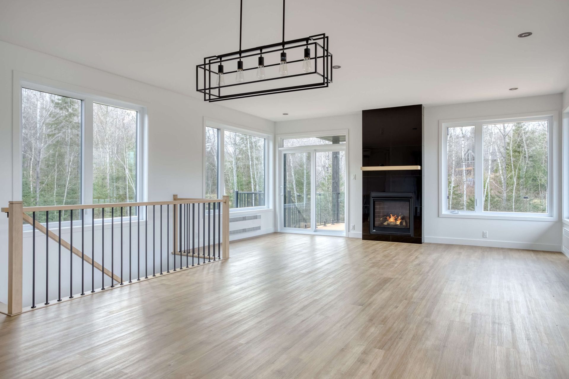 Salon lumineux et vide avec de grandes fenêtres, une cheminée et un escalier. Parquet blond et luminaire noir.