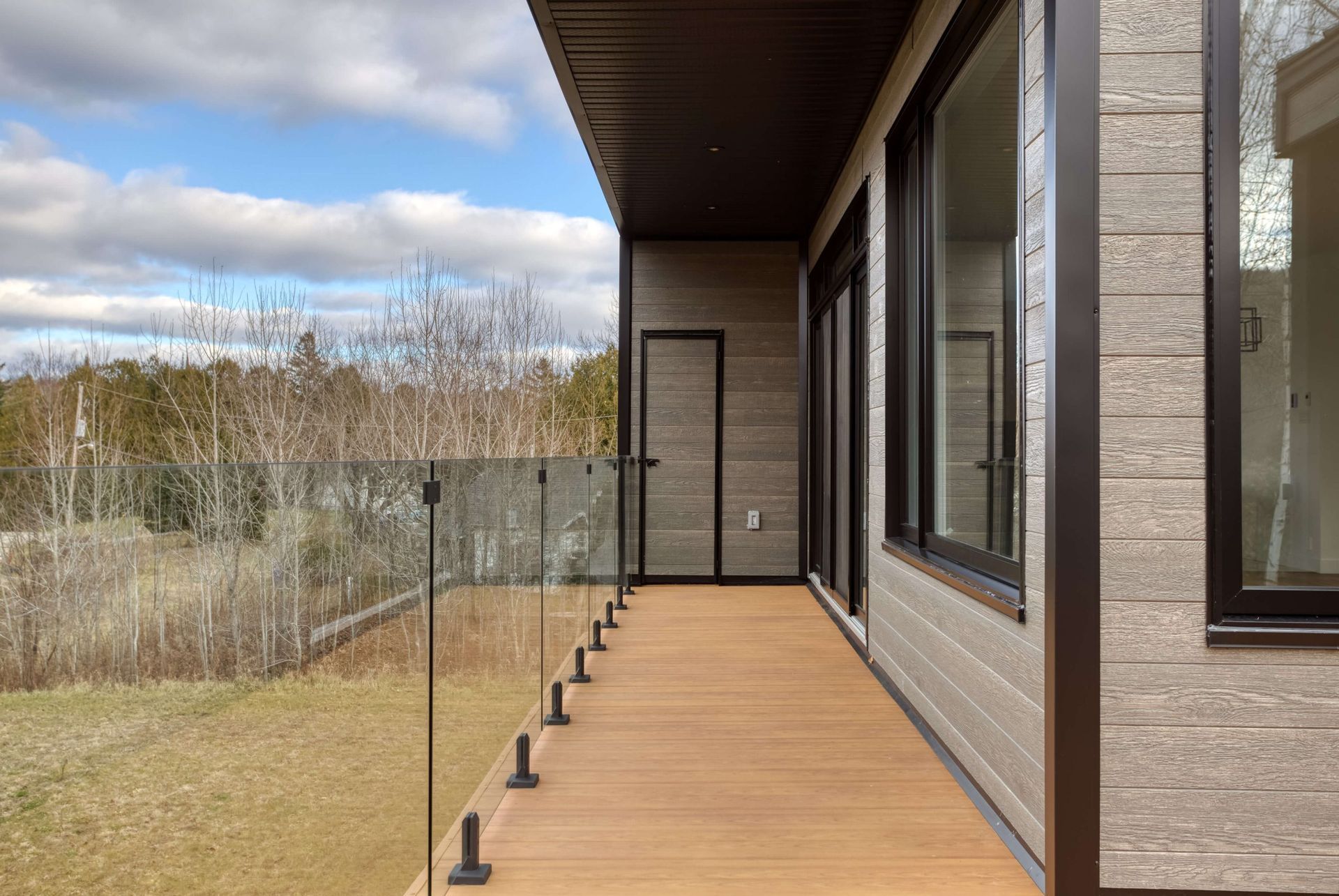 Terrasse en bois avec rambarde en verre et bardage marron. De grandes fenêtres offrent une vue sur les arbres et le ciel.