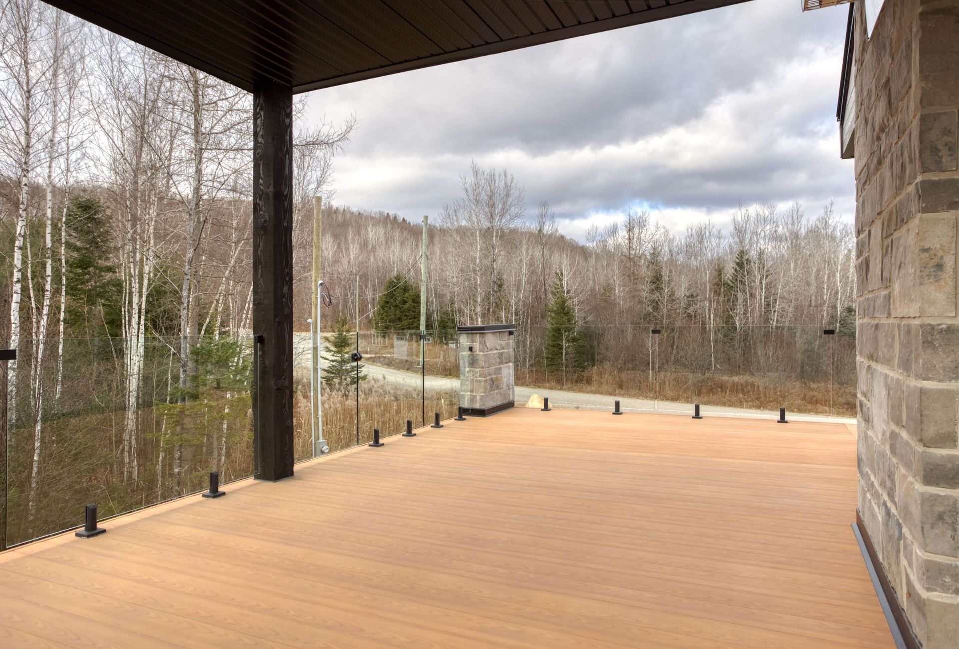 Terrasse en bois avec rambarde en verre, donnant sur une forêt sous un ciel nuageux.