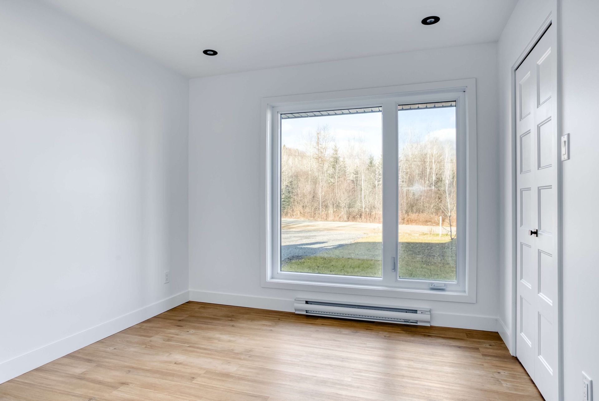 Pièce vide avec murs blancs, fenêtre, parquet et porte blanche.