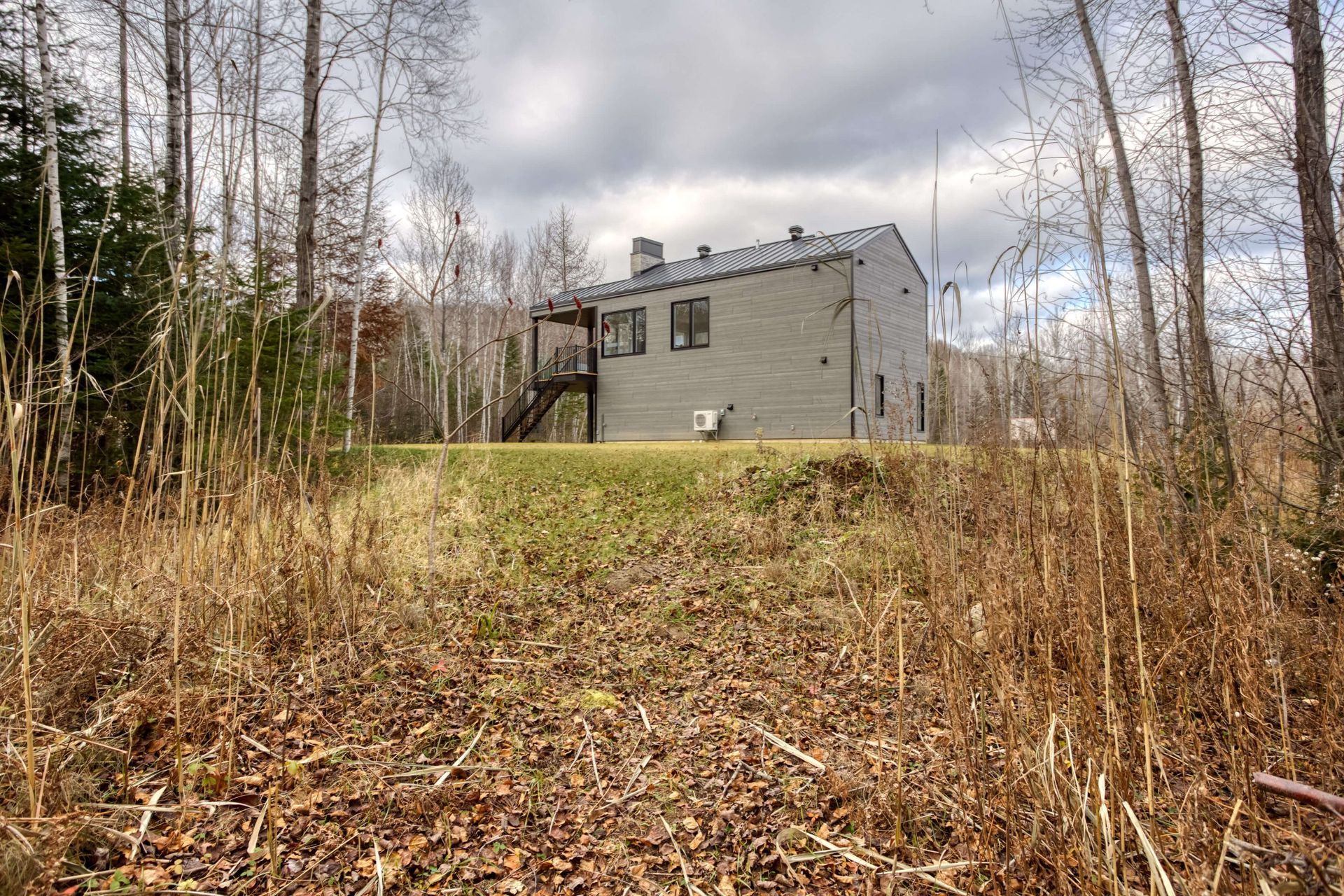 Maison grise moderne sur une colline herbeuse, entourée d'arbres et de hautes herbes brunes sous un ciel nuageux.