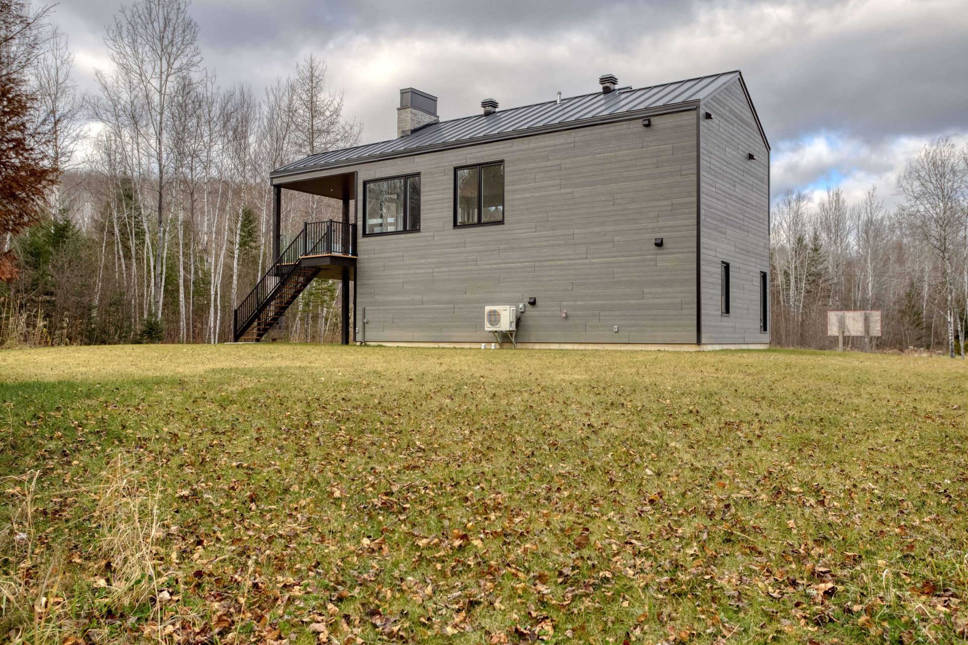 Maison moderne grise avec des touches de noir, située dans un champ herbeux, avec une terrasse et un escalier en bois.