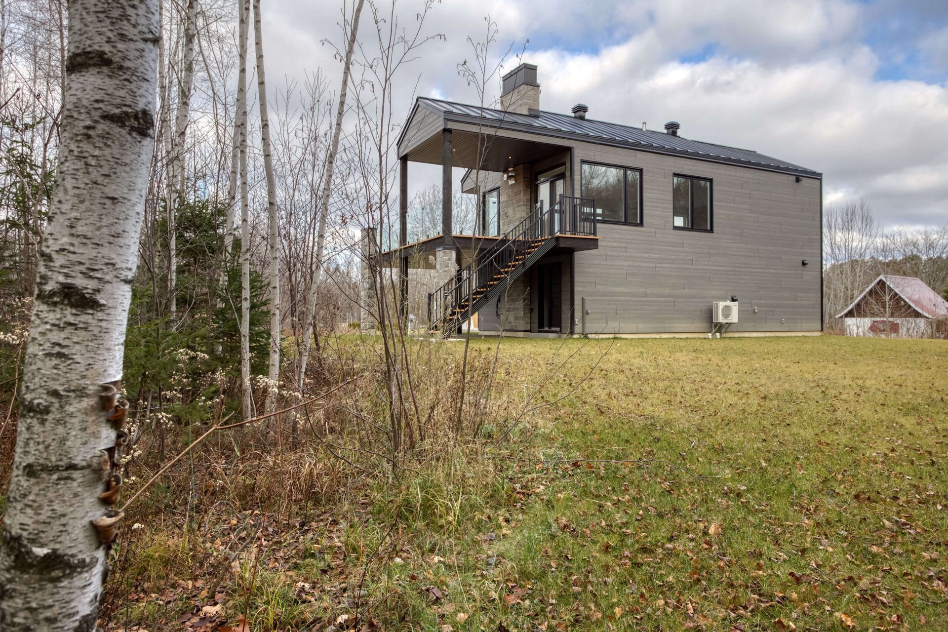 Maison moderne grise avec des touches de noir, entourée d'arbres et d'un champ herbeux.