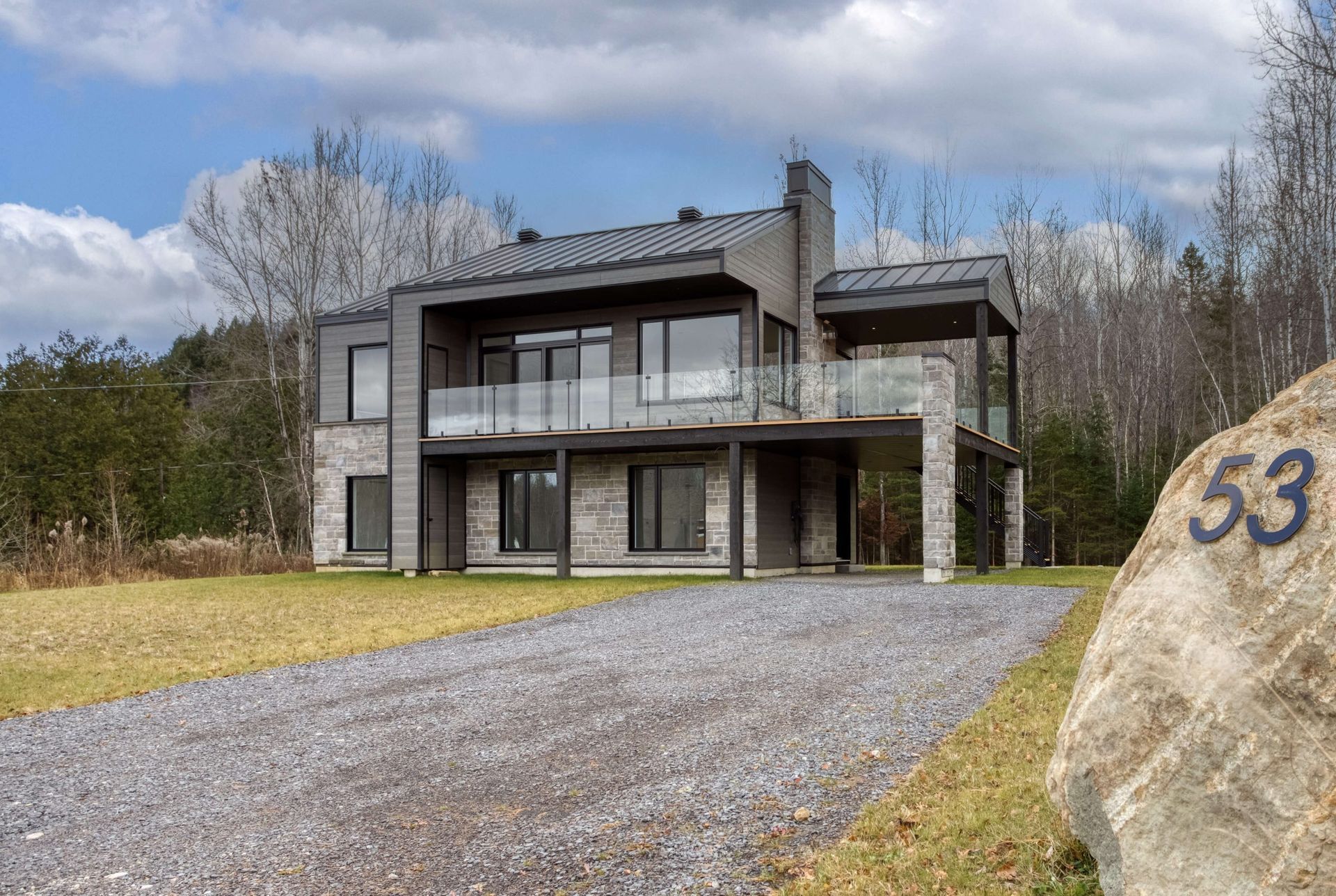 Maison moderne de deux étages avec façade en pierre, balustrades en verre et allée gravillonnée. Numéro « 53 » gravé sur un rocher devant la maison.
