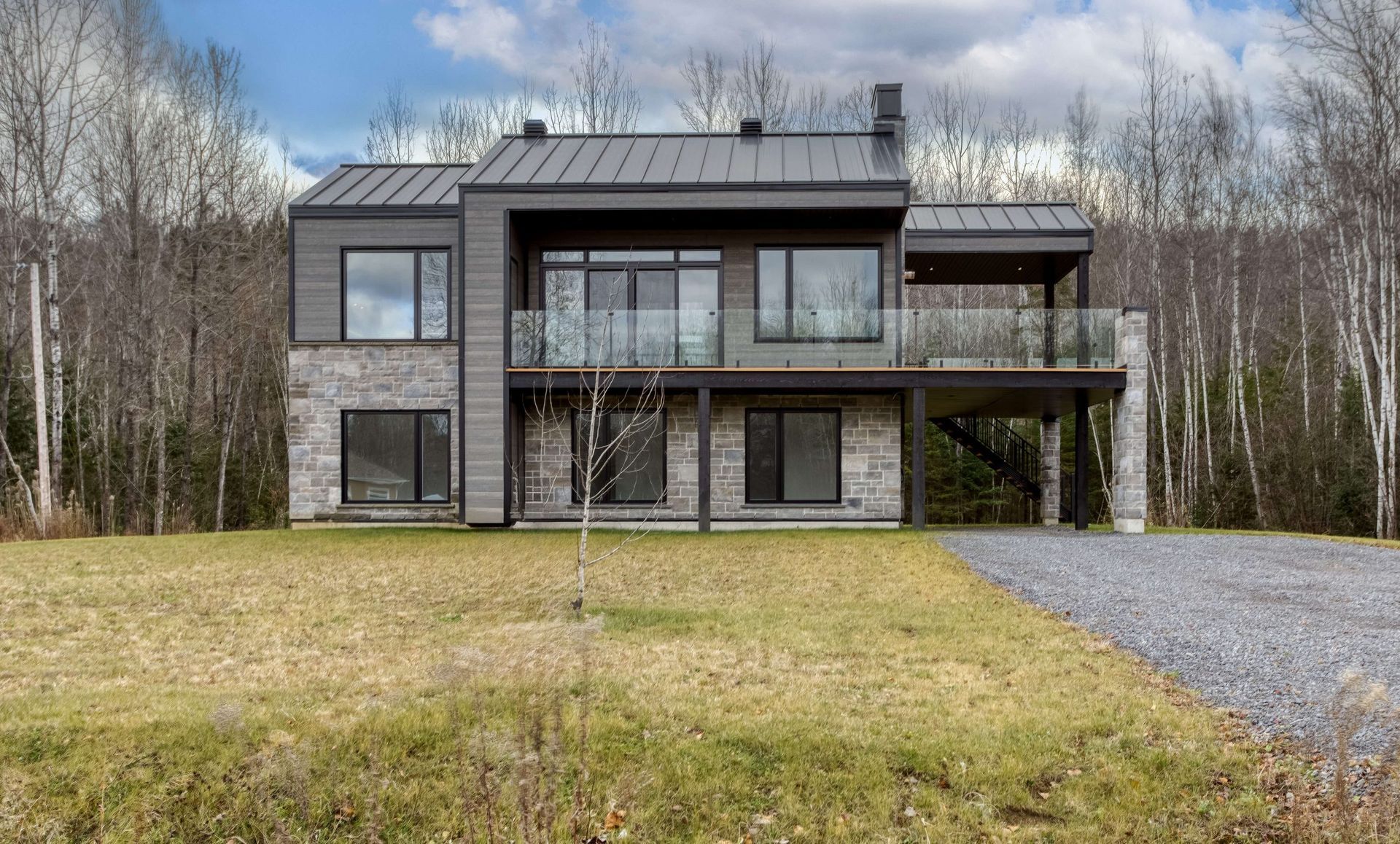 Maison moderne à deux étages avec toit en métal gris, façade en pierre, balcon vitré et allée en gravier.