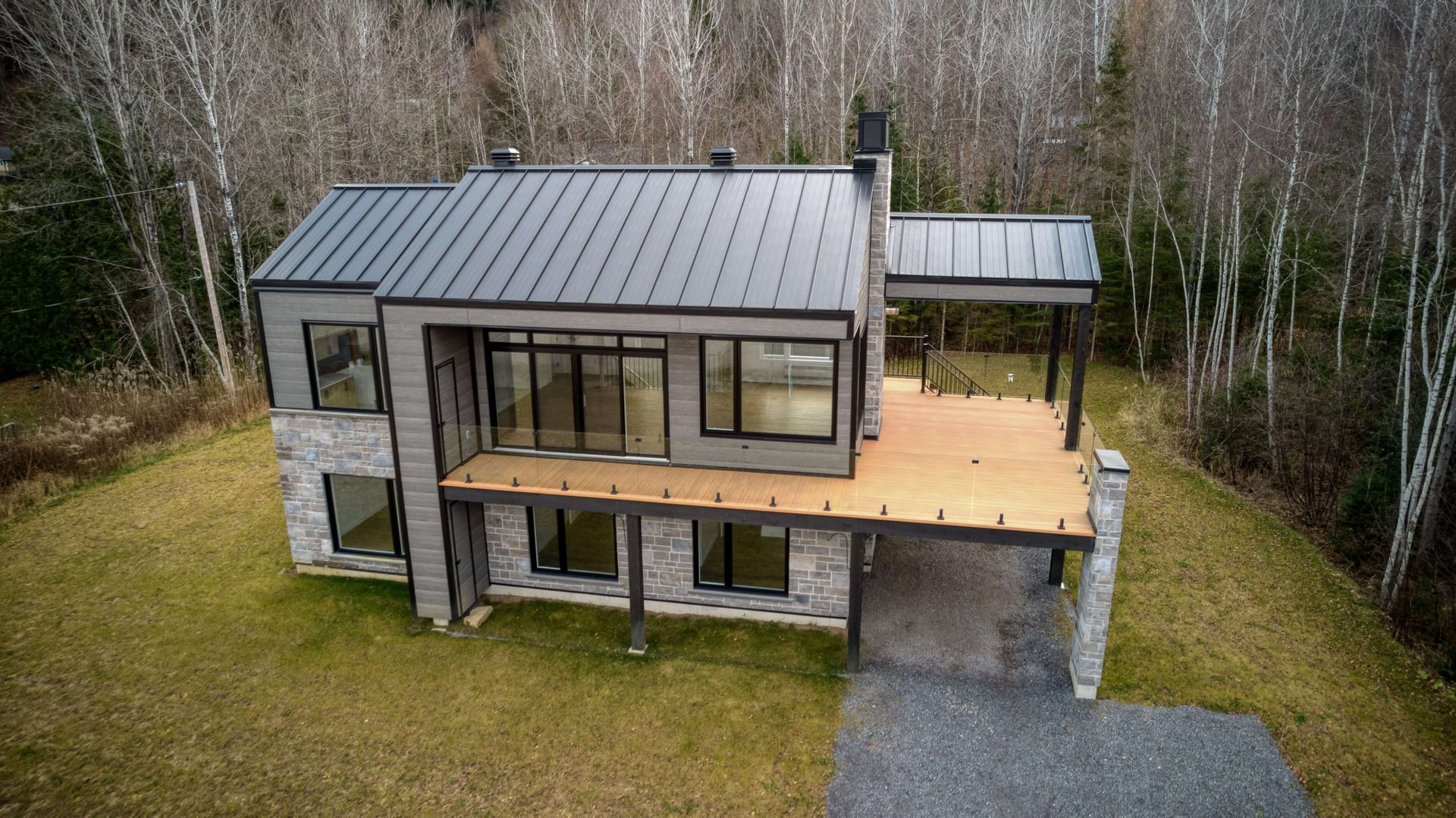 Maison moderne à deux étages avec toit en métal gris, façade en pierre et grandes fenêtres. Terrasse en bois, arrière-plan boisé.
