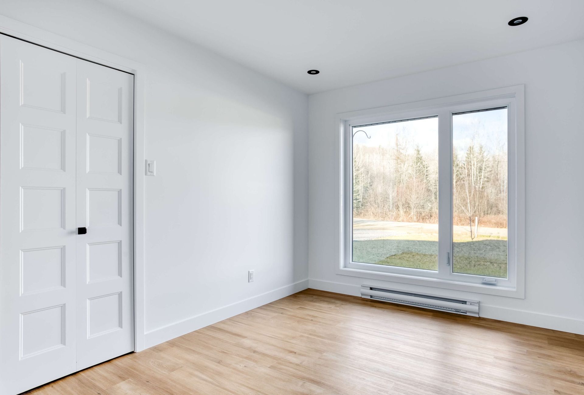 Pièce blanche vide avec porte fermée, fenêtre et parquet.