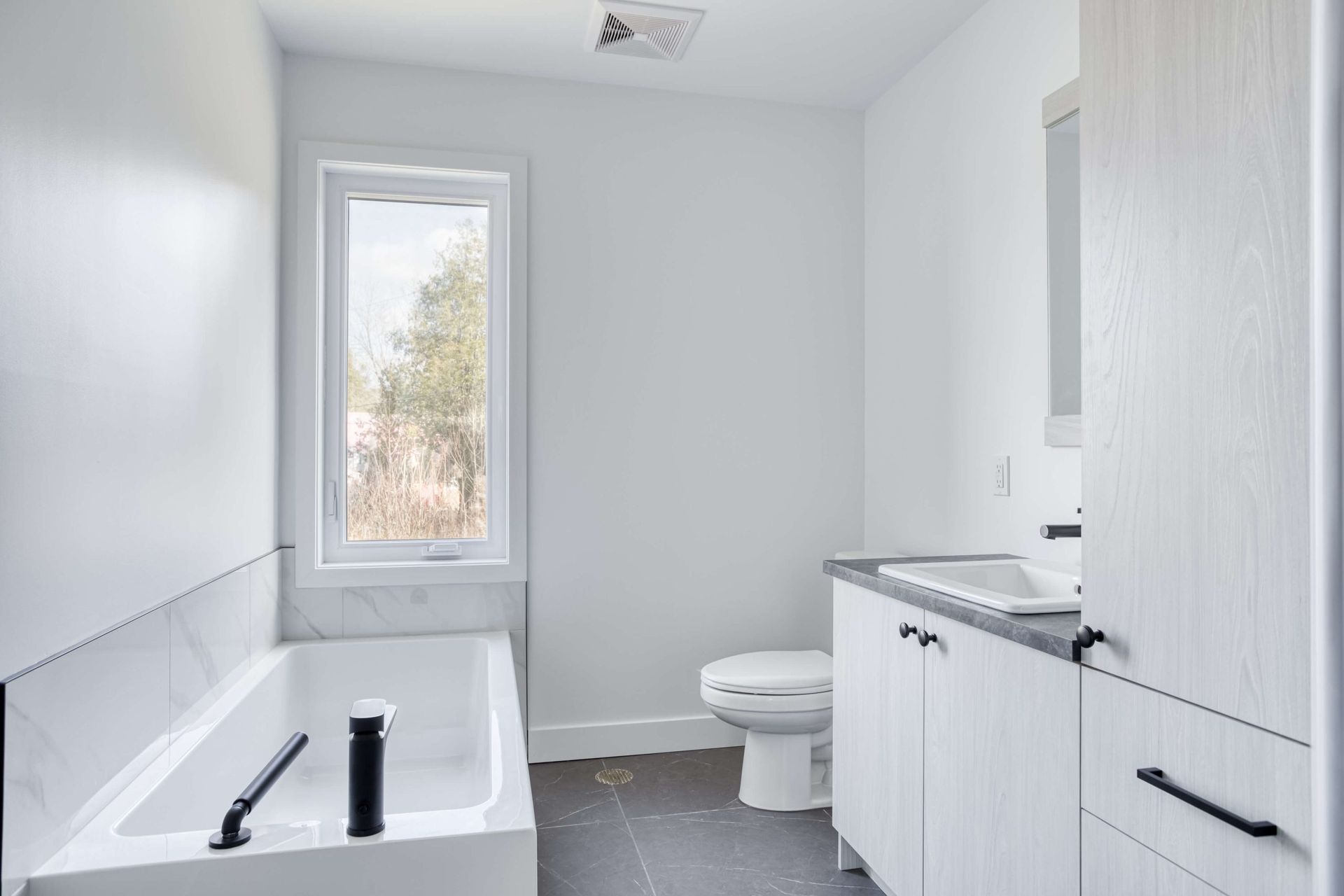 Salle de bains blanche avec baignoire, toilettes et meuble-lavabo près d'une fenêtre.