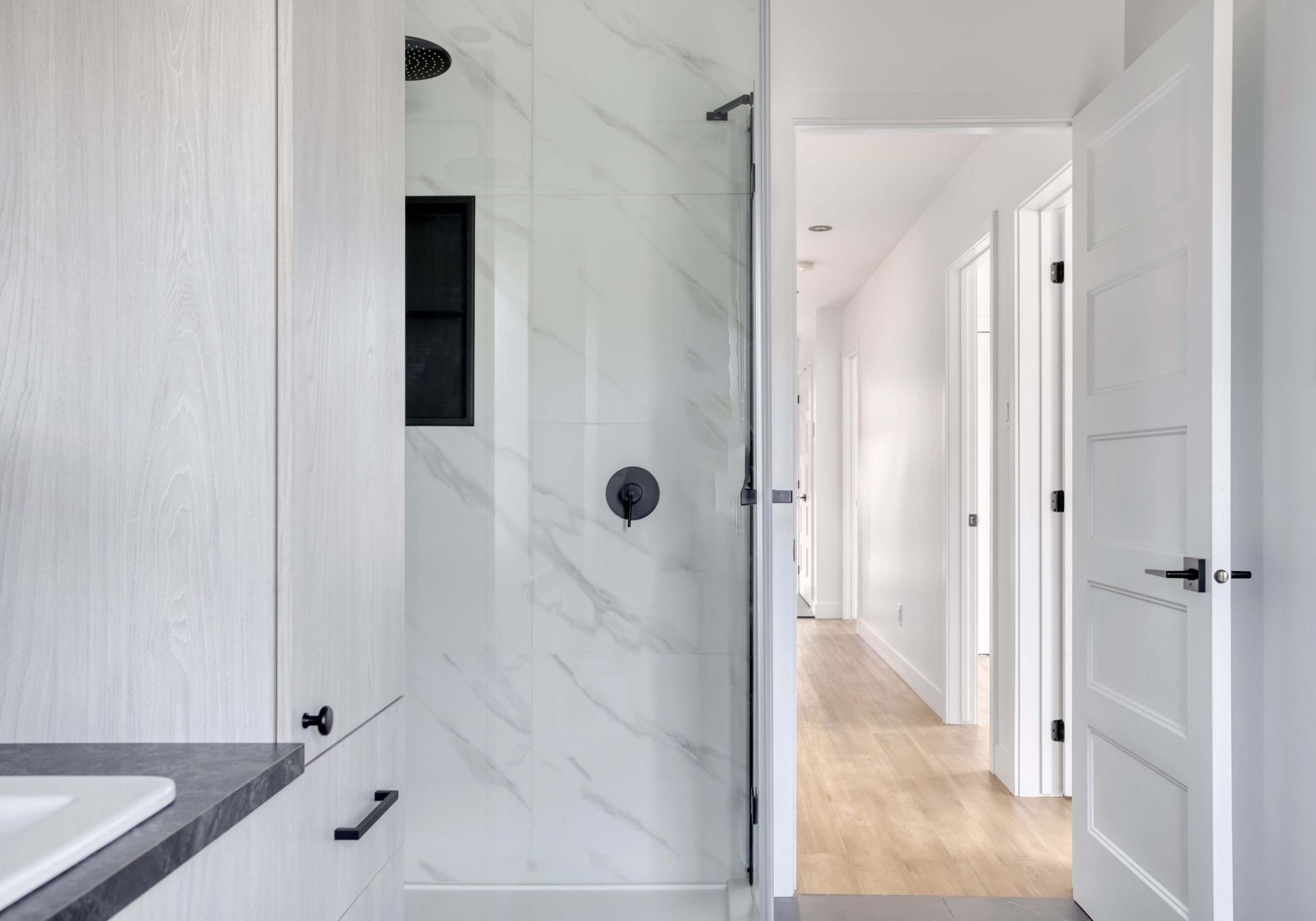 Salle de bains avec meubles blancs, douche en marbre, porte ouverte sur le couloir.