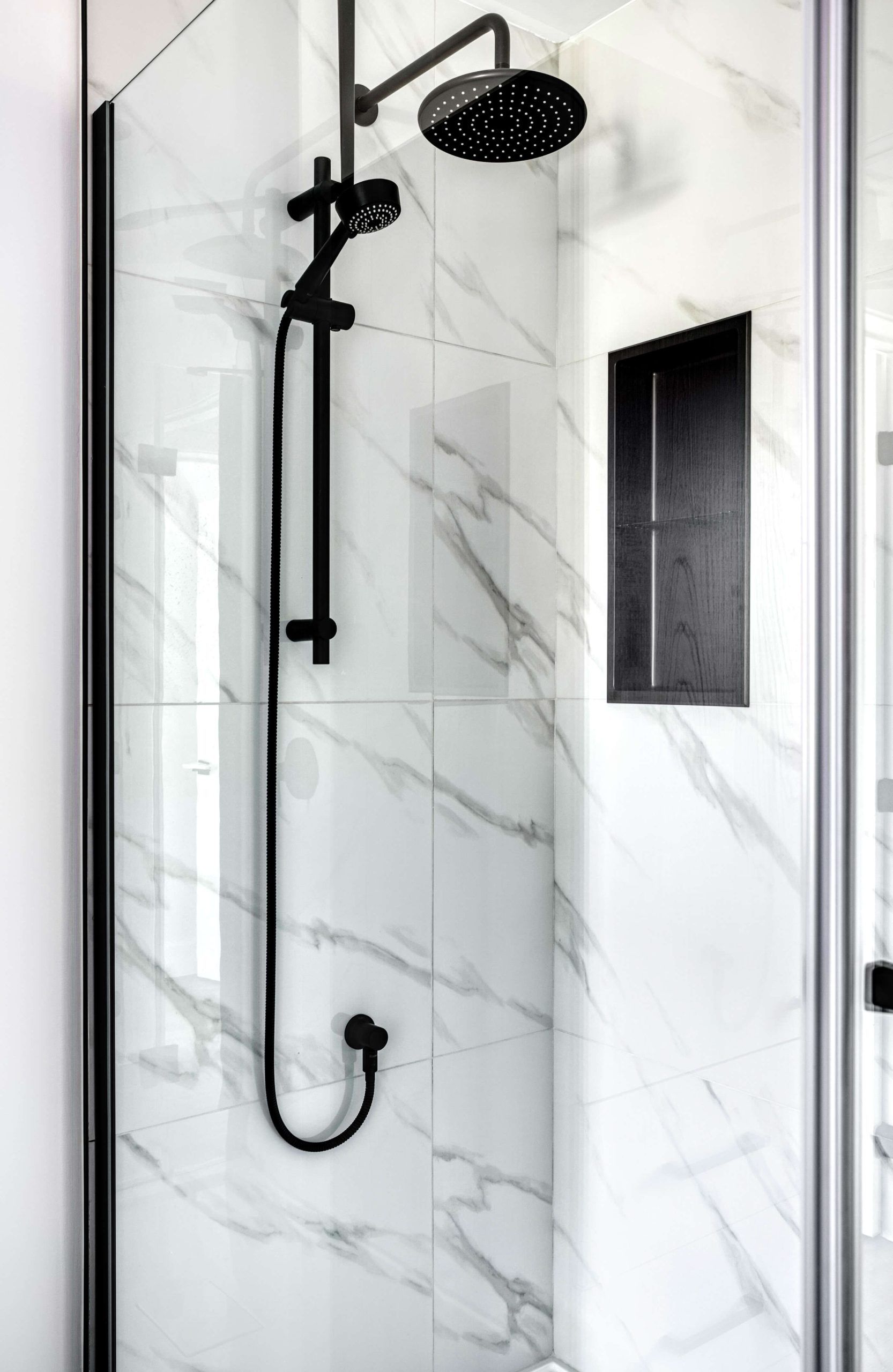 Robinetterie de douche noire contre un mur carrelé de marbre blanc, avec une étagère intégrée.