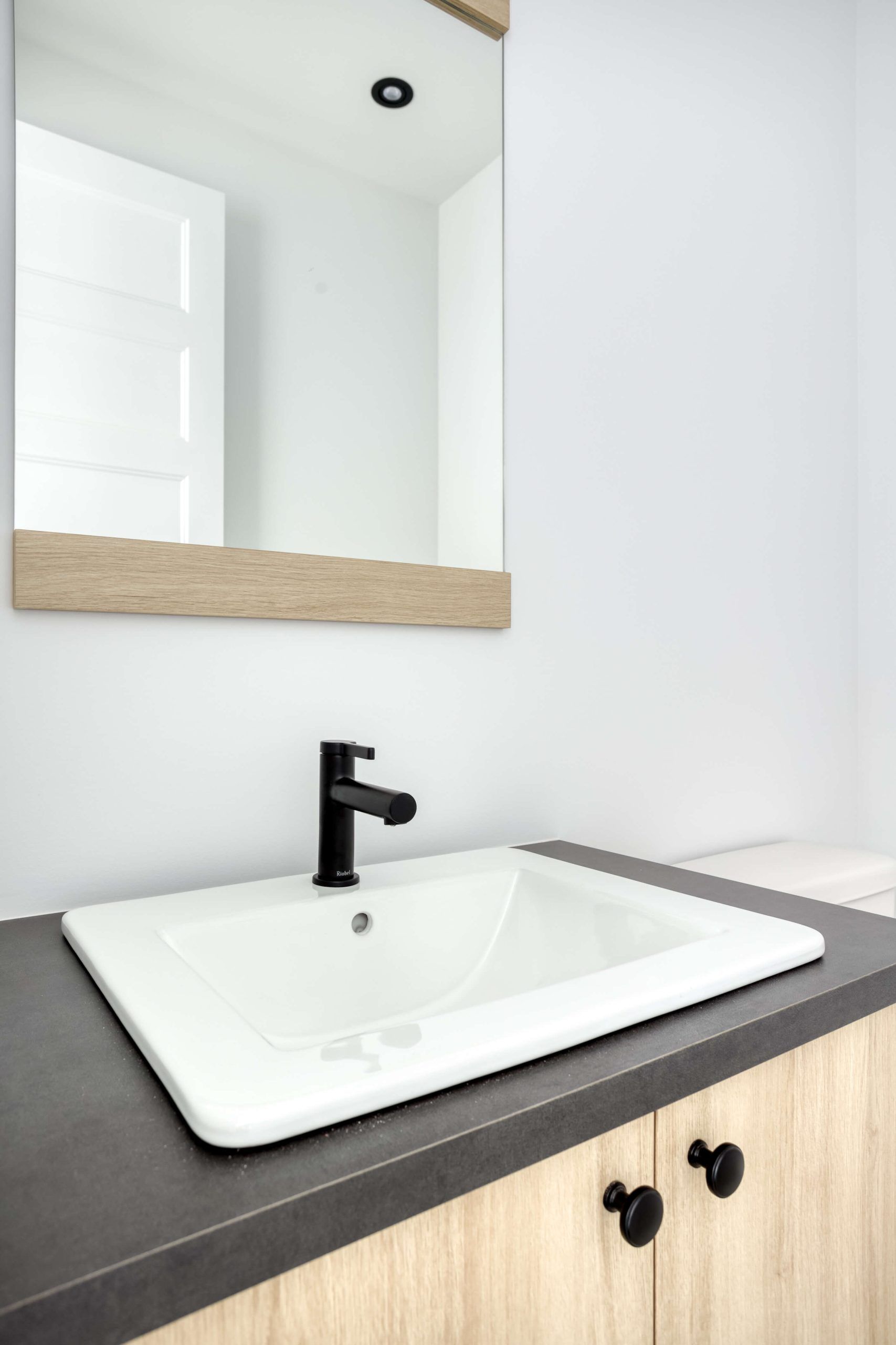 Lavabo de salle de bains avec robinet noir, vasque blanche, plan de travail gris et meuble en bois. Miroir au-dessus.
