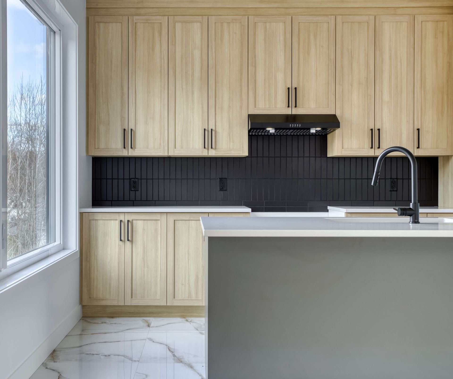 Cuisine moderne avec armoires en bois clair, crédence noire, îlot et fenêtre.