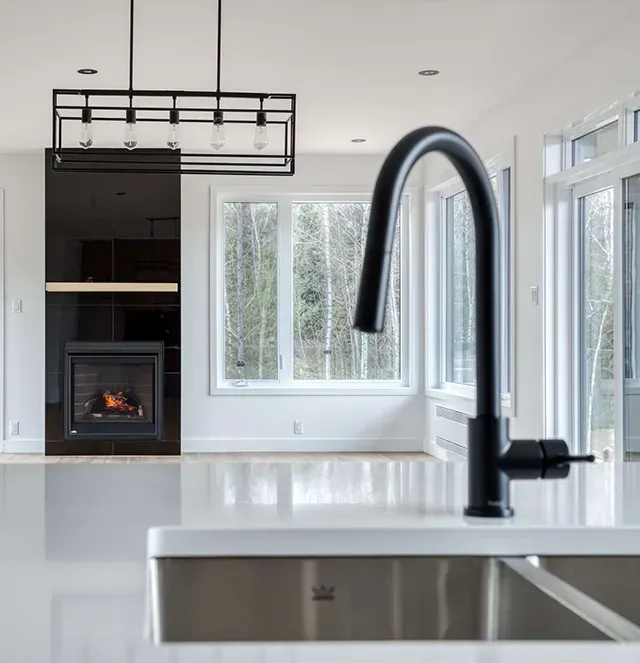 Robinet de cuisine noir devant un plan de travail blanc et un salon flou avec une cheminée et des fenêtres.