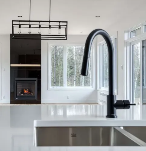 Robinet de cuisine noir devant un plan de travail blanc et un salon flou avec une cheminée et des fenêtres.