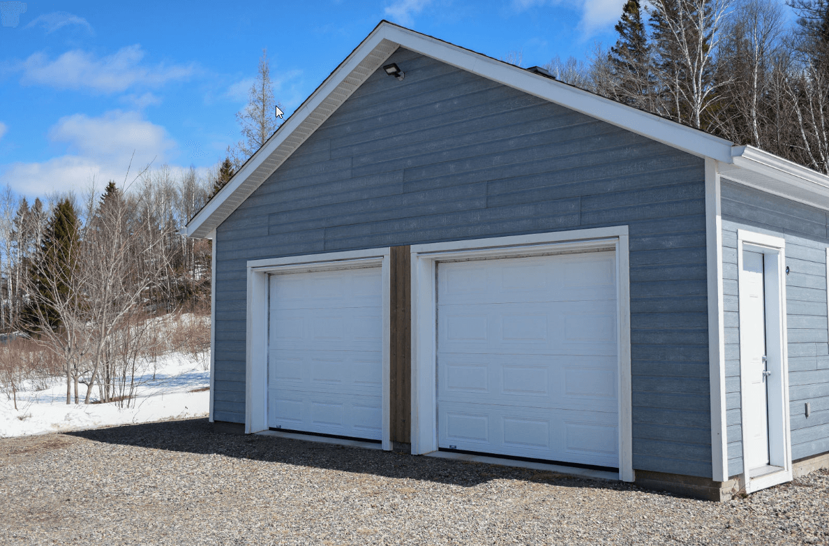 Garage bleu avec deux portes blanches, allée de gravier et neige en arrière-plan sous un ciel bleu.