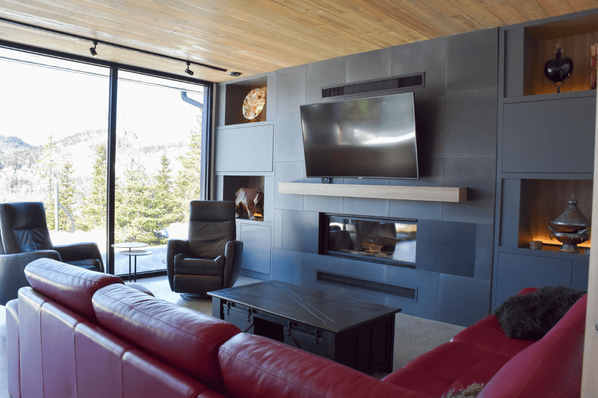 Salon avec canapé rouge, cheminée, grandes fenêtres et mur carrelé gris avec une télévision.