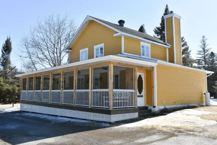 Maison jaune à deux étages avec une véranda qui l'entoure, située dans un paysage hivernal enneigé et parsemé d'arbres dénudés.