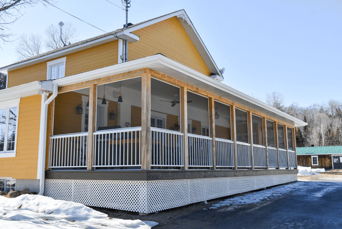 Maison jaune avec véranda grillagée, balustrade blanche et plinthe en treillis ; neige au sol.