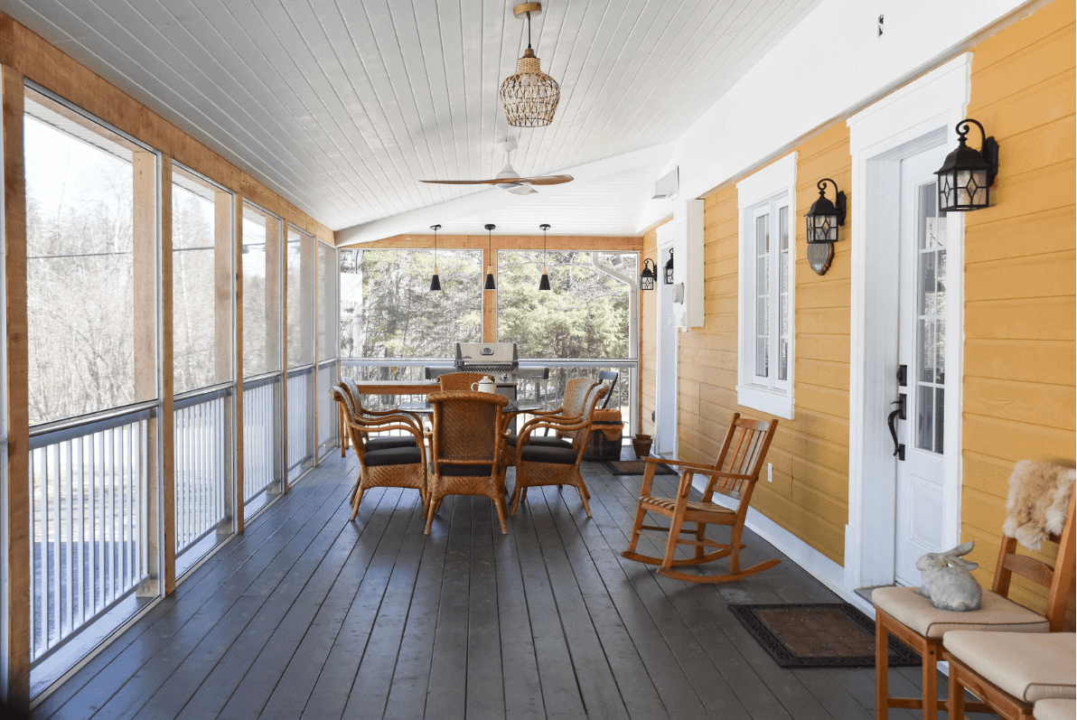 Véranda jaune avec sol foncé, meubles en osier, boiseries blanches et fenêtres moustiquaires.