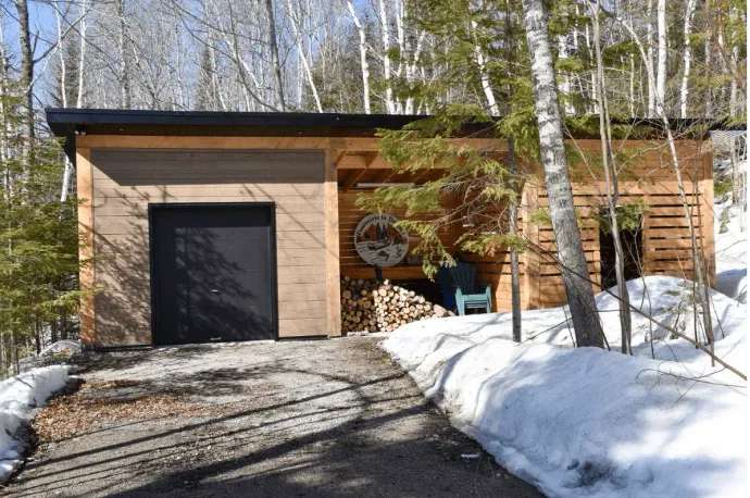 Un abri de jardin avec une porte noire, du bois de chauffage et une allée enneigée dans une zone boisée.