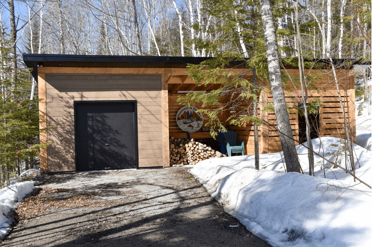 Abri en bois et en pierre avec porte noire, bois de chauffage et neige.