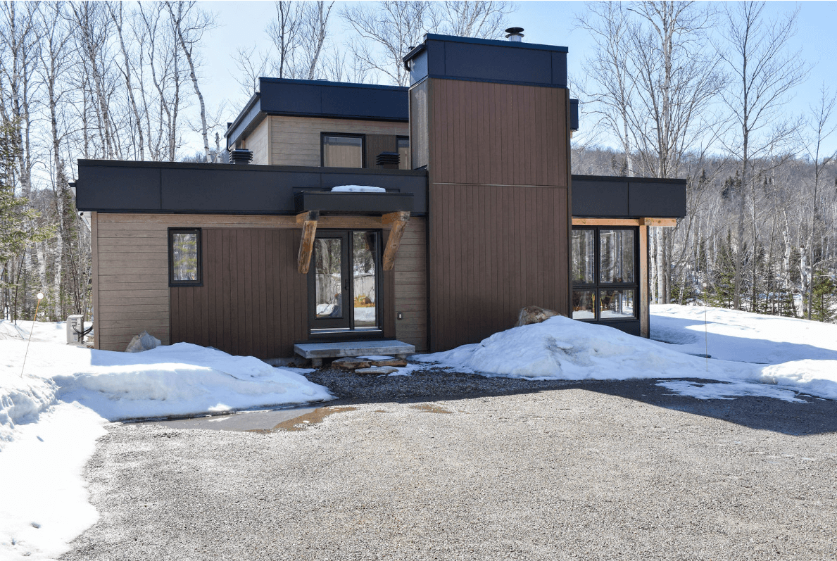 Maison moderne brune et beige avec un toit plat, entourée de neige et d'arbres.