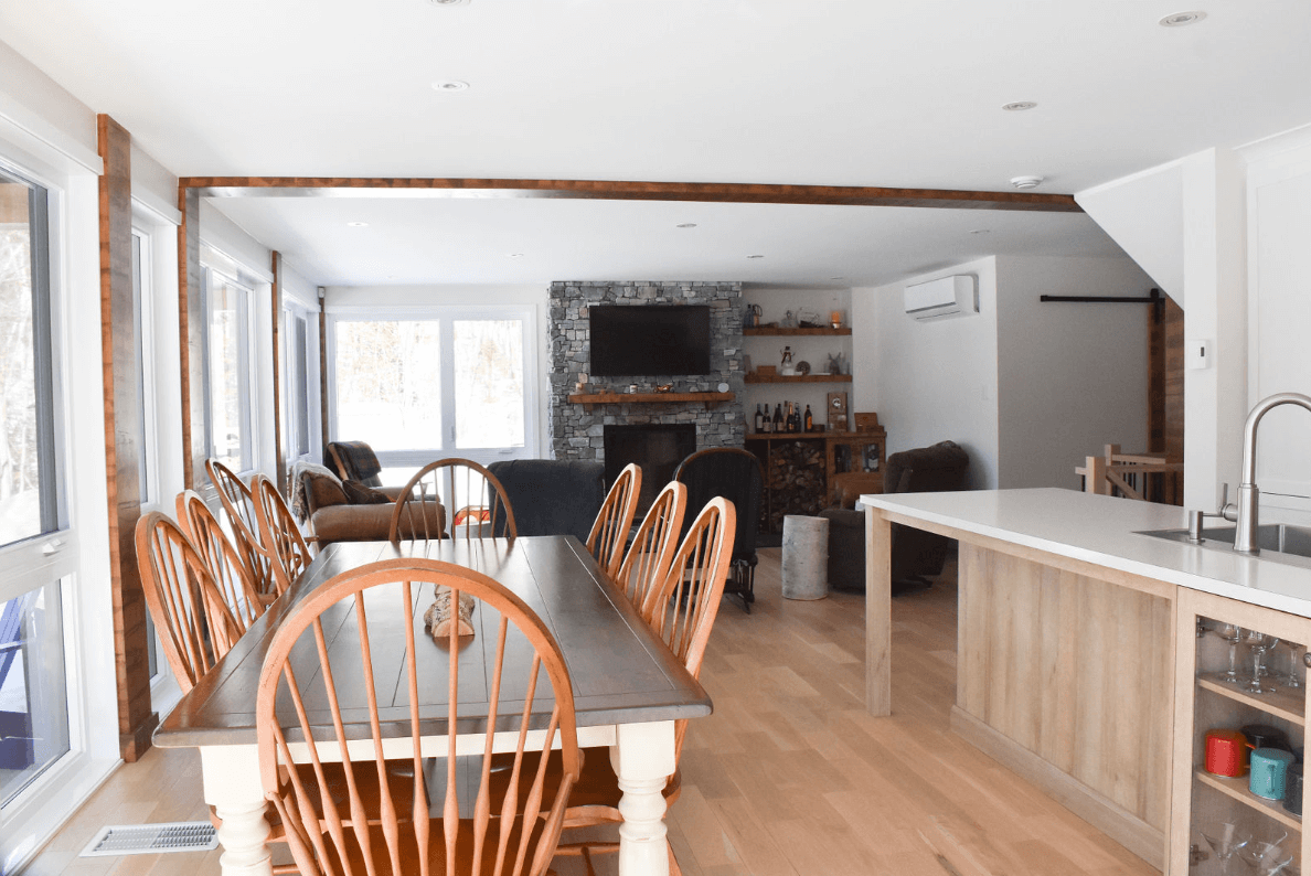 Salle à manger avec grande table et chaises en bois, ouverte sur le salon avec cheminée et îlot de cuisine.