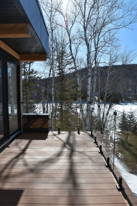 Terrasse en bois avec rambarde en verre, donnant sur une forêt enneigée. Le soleil projette des ombres sur la terrasse.