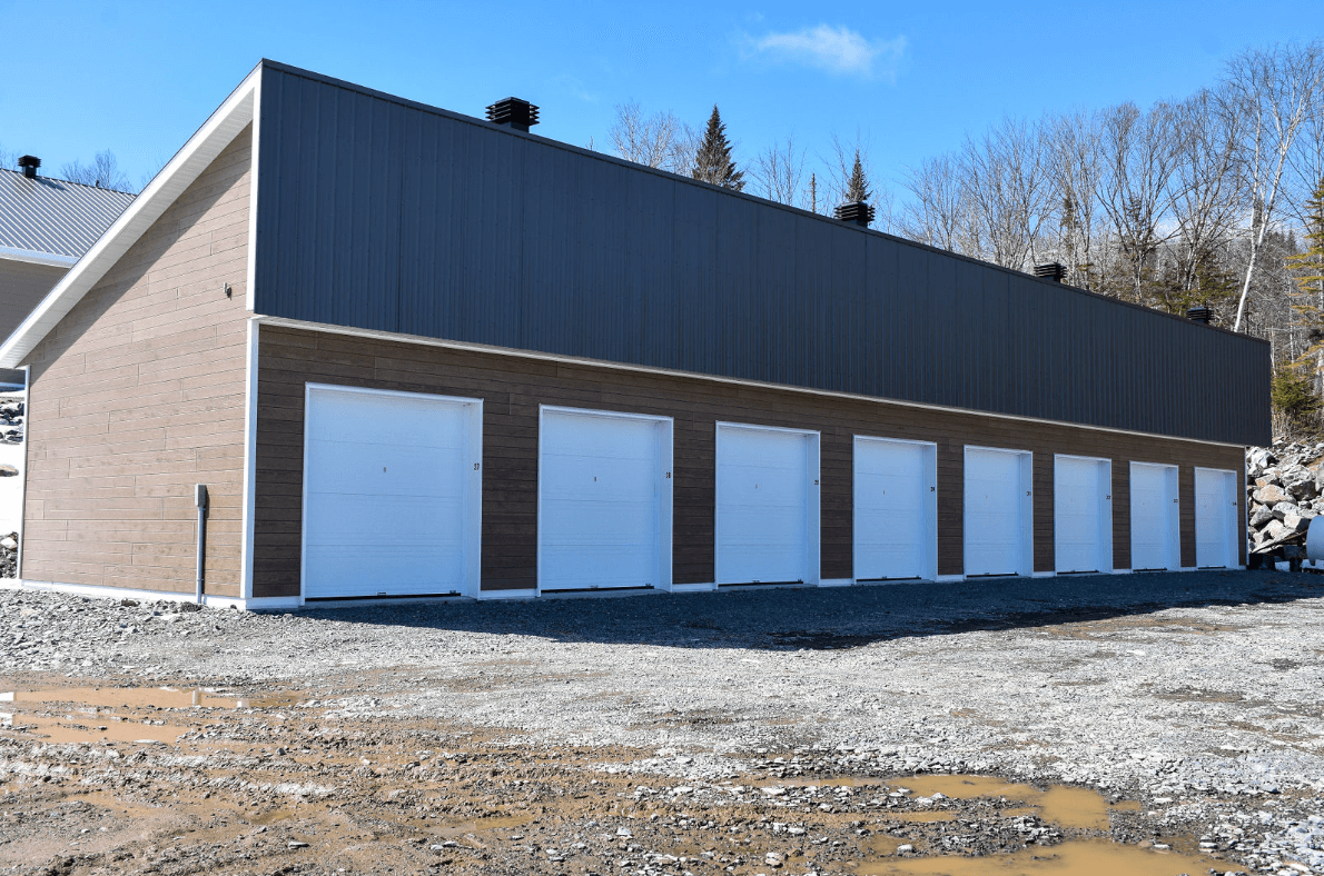 Une rangée de portes de garage blanches sur un bâtiment en briques avec un toit en métal sombre, sous un ciel bleu.