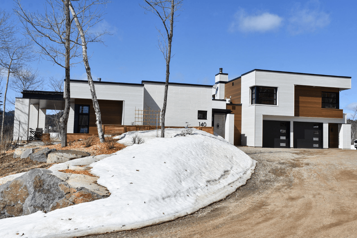 Maison moderne blanche et en bois, avec une allée enneigée et un ciel bleu.