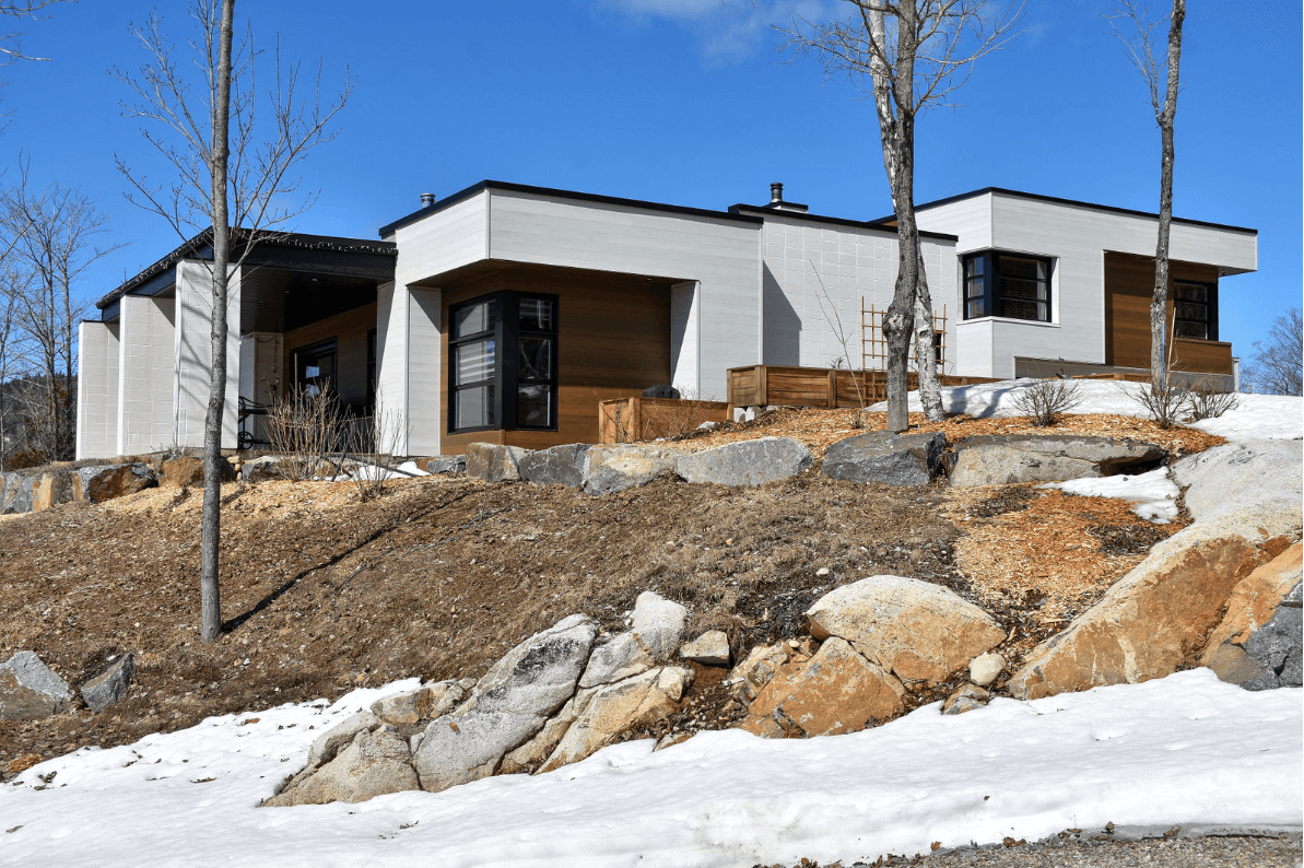 Maison moderne avec façade en stuc blanc et éléments en bois, sur un flanc de colline rocailleux et enneigé.