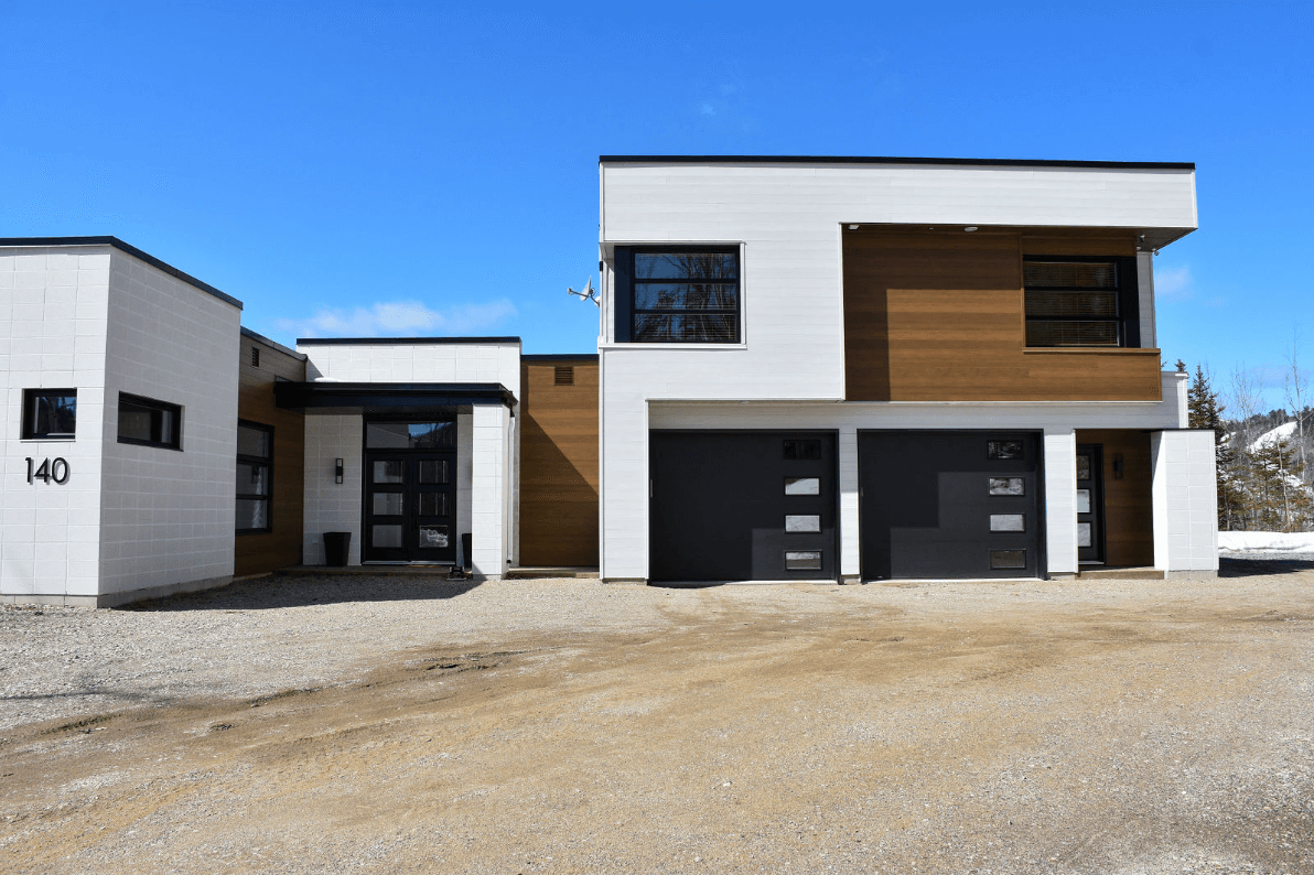 Maison moderne aux murs blancs, bardage en bois brun, portes de garage et fenêtres noires ; allée en gravier.