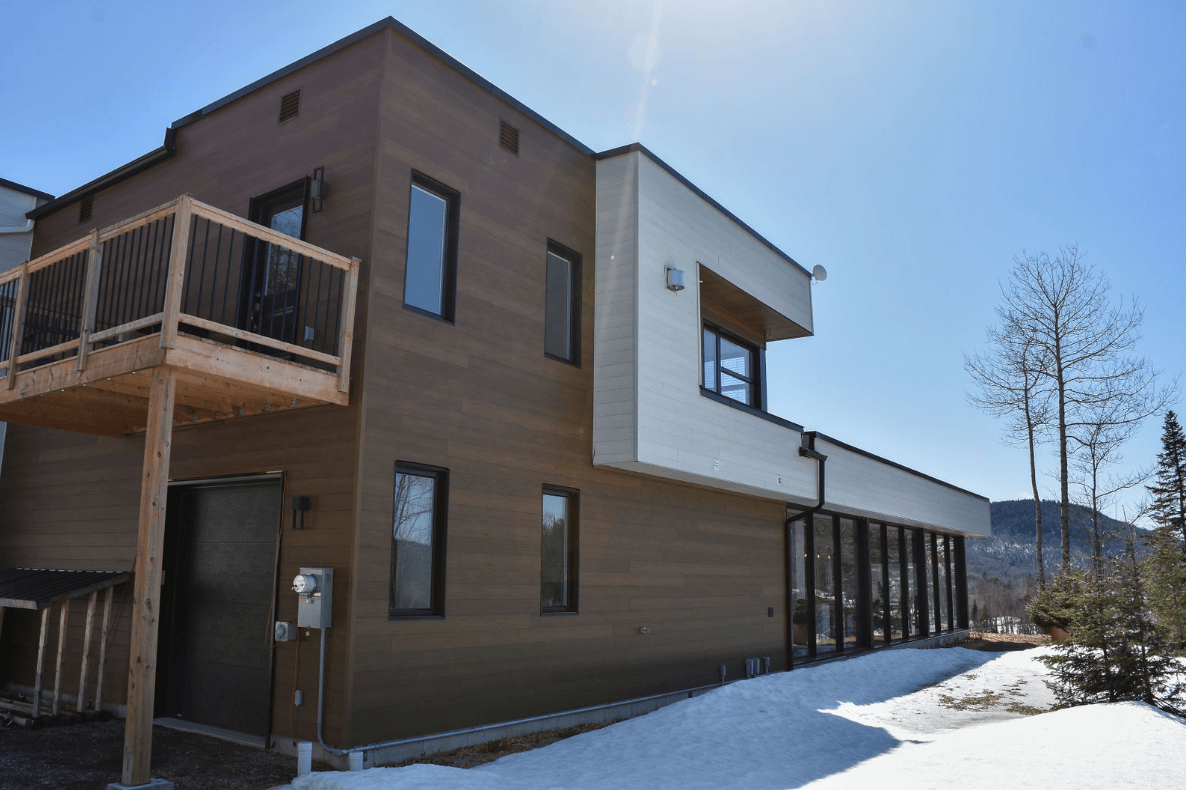Maison moderne à deux étages avec façade marron et blanche, terrasse en bois, neige au sol et ciel ensoleillé.