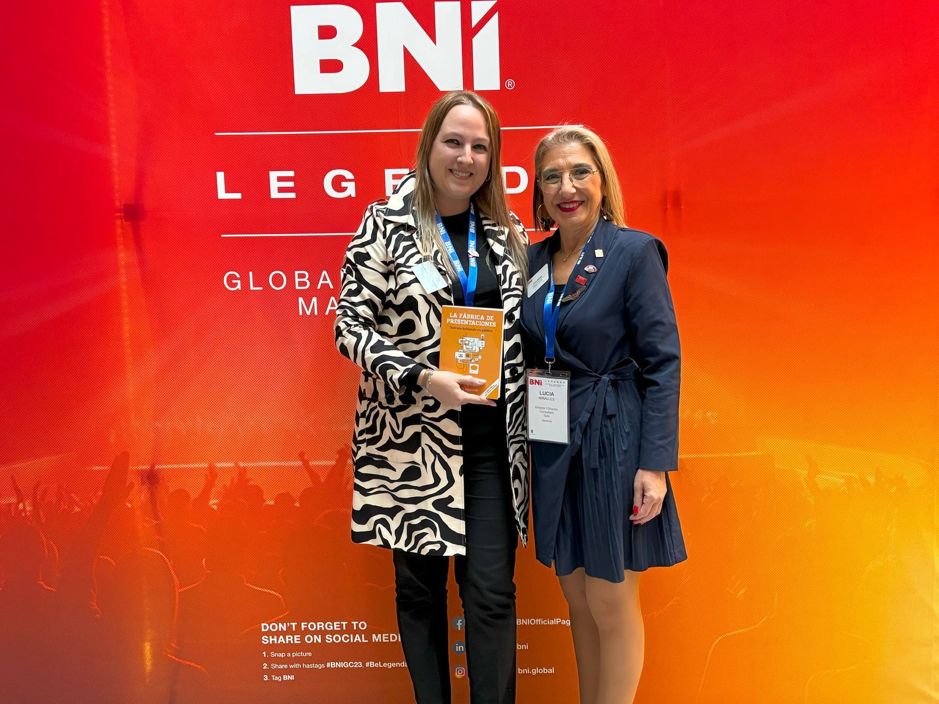 Dos mujeres posando en un evento de BNI. Una sostiene un libro y ambas sonríen. Fondo rojo y naranja.