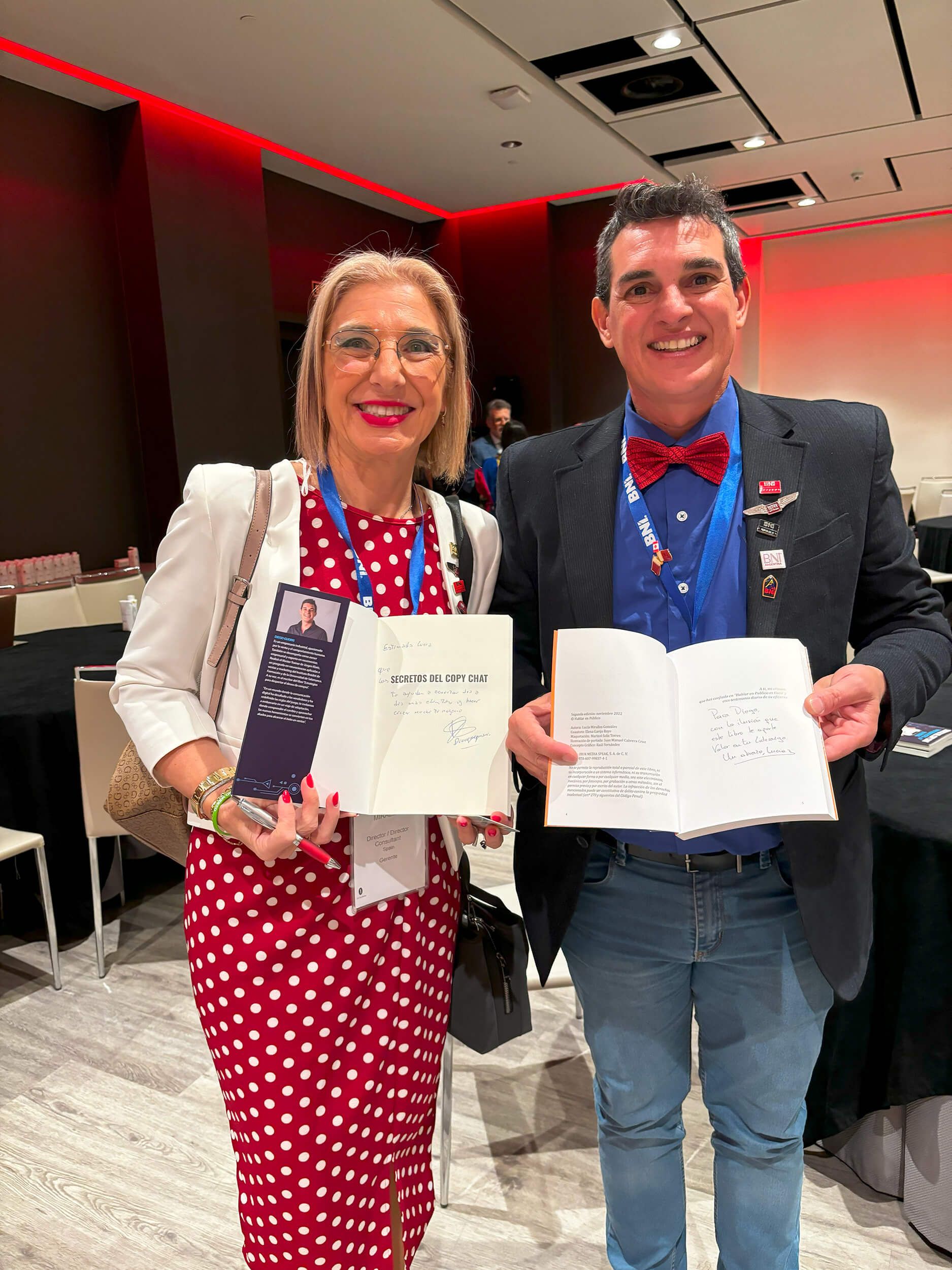 Dos personas sonriendo, con papeles en la mano, en una conferencia. Una mujer con lunares rojos y un hombre con vaqueros y pajarita roja.