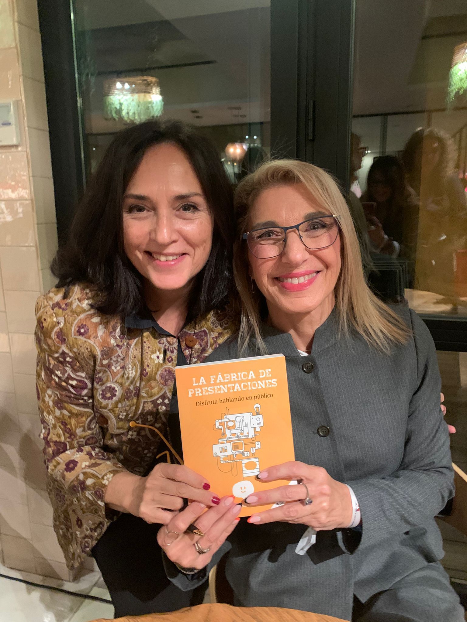 Mujer con lunares y hombre con libro sonriendo juntos en el interior.