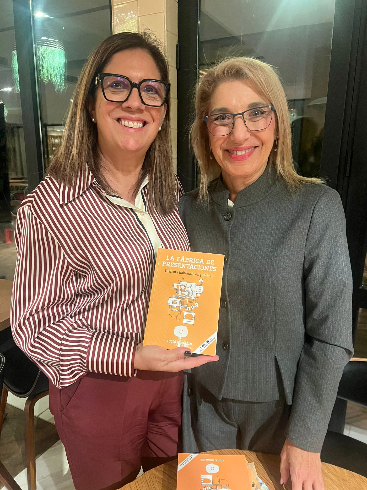 Dos personas sonriendo, con papeles en la mano, en una conferencia. Una mujer con lunares rojos y un hombre con vaqueros y pajarita roja.