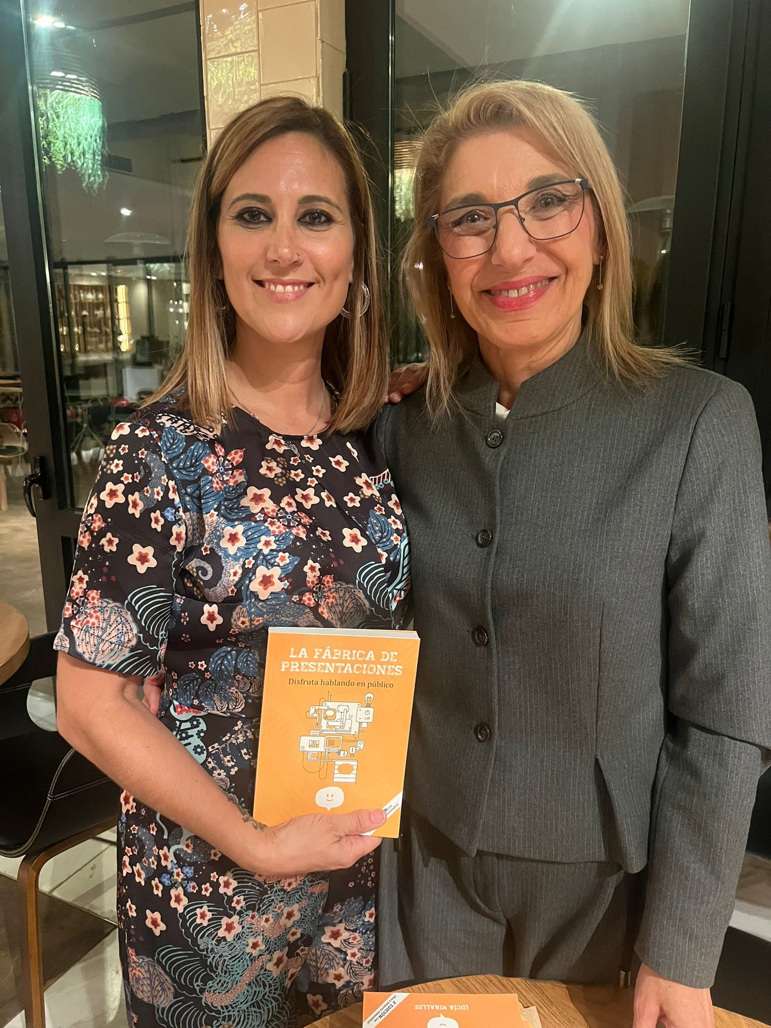 Dos personas sonriendo, posando juntas en un interior. Una mujer con un vestido rojo de lunares y un hombre con traje sosteniendo un libro.