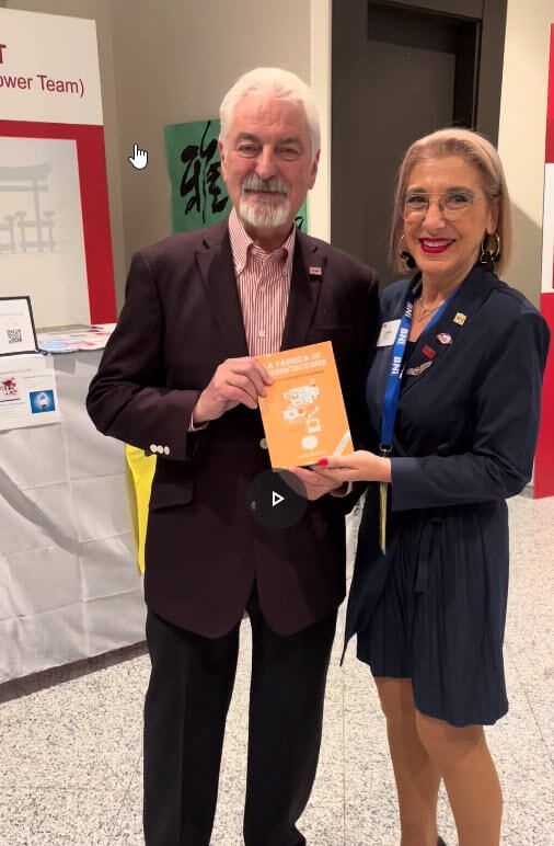 Un hombre y una mujer sonríen, sosteniendo un libro. Están de pie frente a una mesa de exposición.