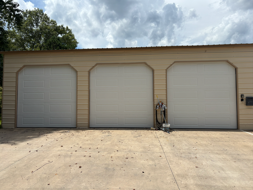 Three white garage doors on a beige metal building with a concrete driveway.