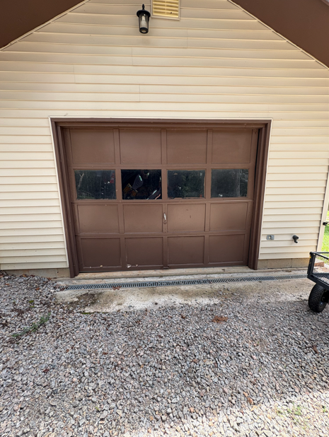 Brown garage door with four window panes, tan siding, gravel ground.
