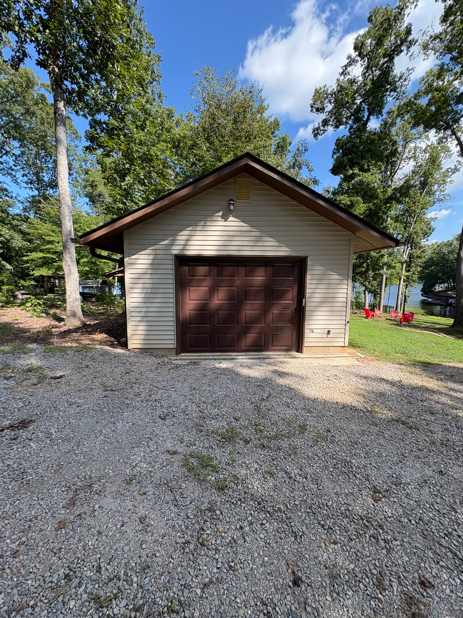 A garage with a brown door and tan siding sits on a gravel driveway, with trees and water in the background.