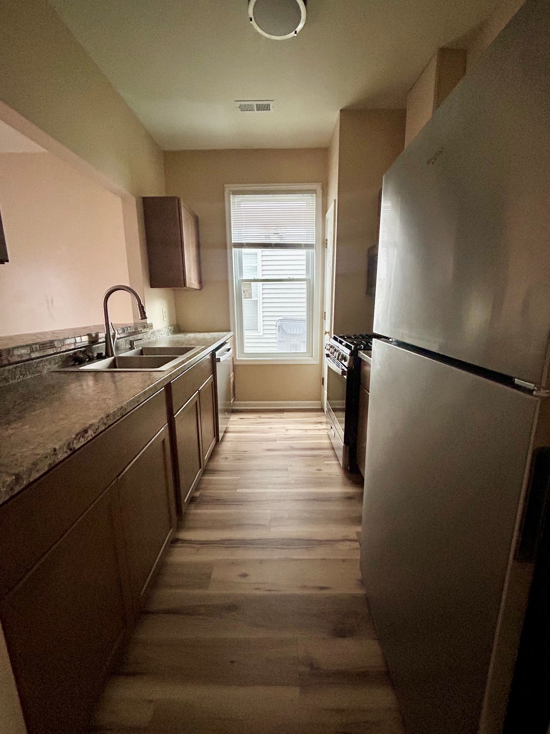 Long, narrow kitchen with wood cabinets, light countertops, and a refrigerator. Window at end.