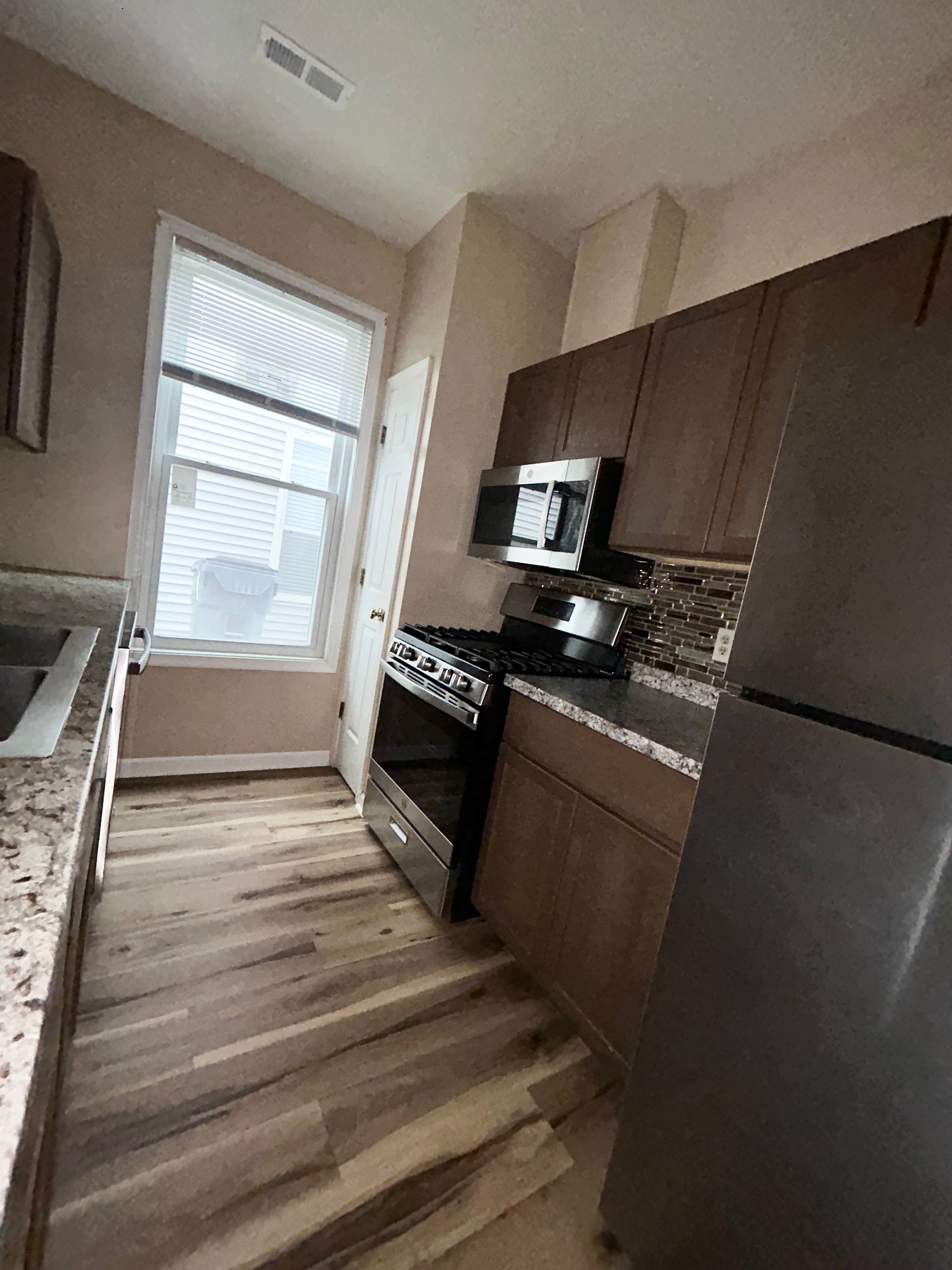 Kitchen with dark brown cabinets, stainless steel appliances, and wood-look flooring. Window with blinds visible.
