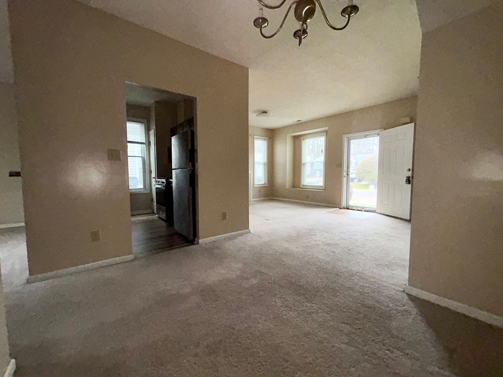 Interior of a room with carpet, open doorway to kitchen with black fridge, and another door to outside.