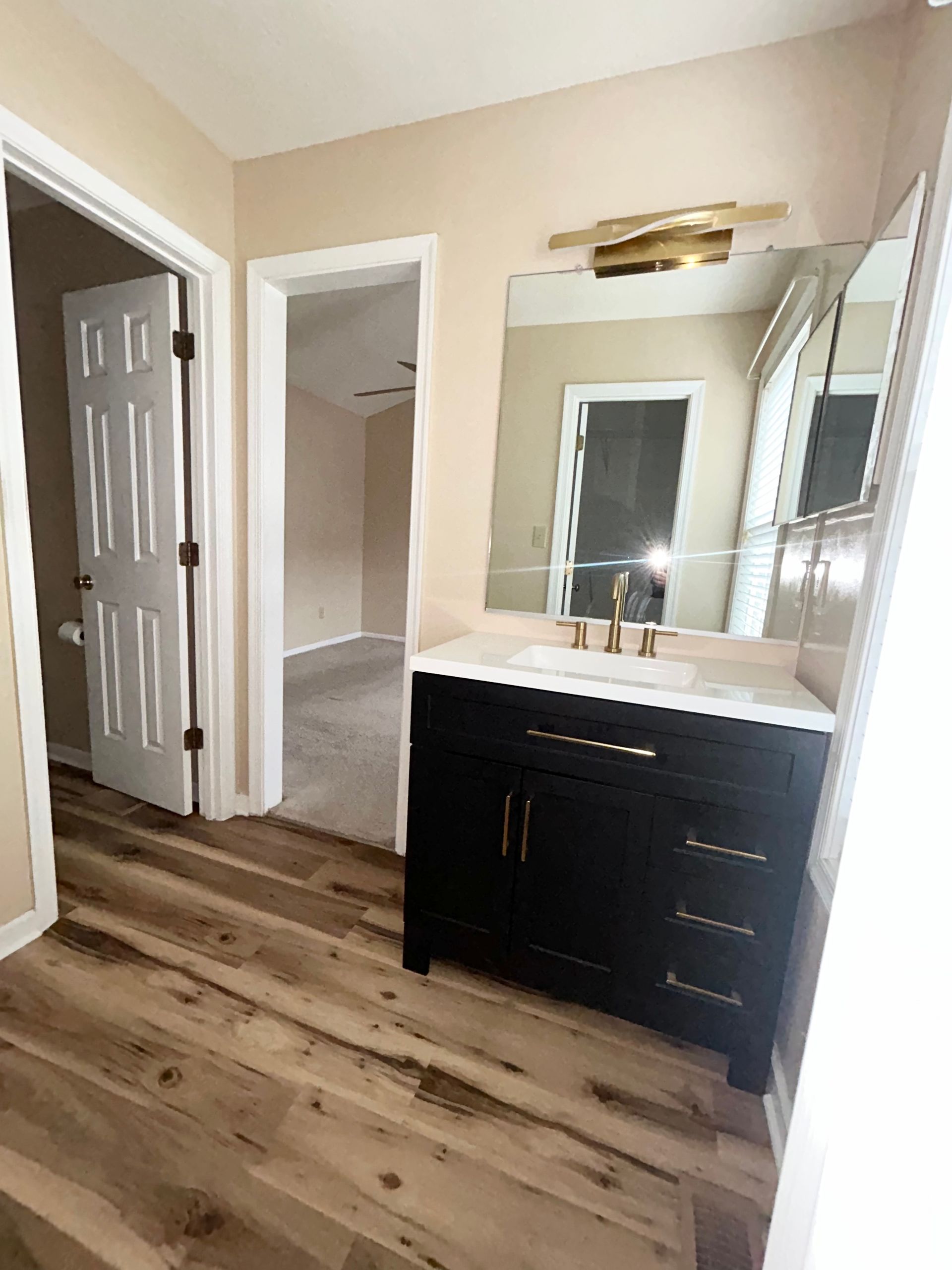 Bathroom with a black vanity, gold fixtures, and a large mirror. Brown wood-look flooring. Two doorways.